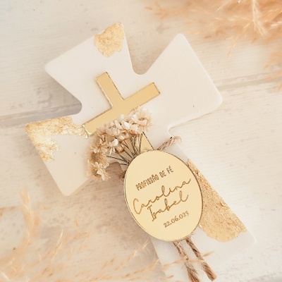 Cruz branca e dourada com flores secas e etiqueta personalizada sobre mesa clara