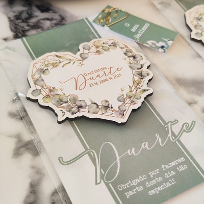 Tag de batizado em formato de coração com design floral e texto, sobre cartão verde com texto e embalagem transparente