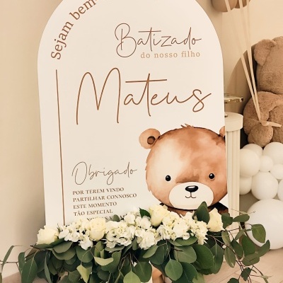 Placa decorativa para batizado com texto e desenho de urso, flores brancas e balões castanho e branco.