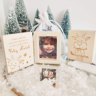 Molduras de madeira clara com desenhos de rena e texto em português, neve artificial e pinheiros nevados ao fundo