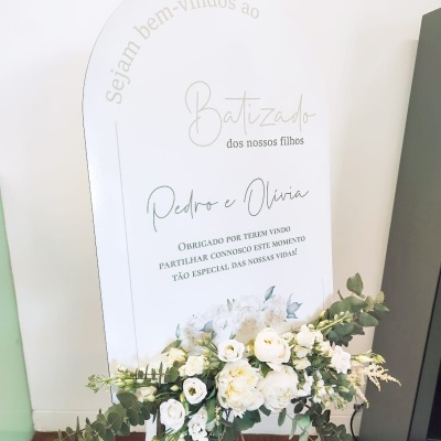 Placa branca com texto dourado e preto sobre batizado com flores brancas e verdes na base