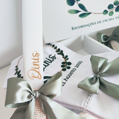 Vela decorativa branca com nome Dinis e laço verde, ao lado de pano branco bordado com texto e ilustração de folhas