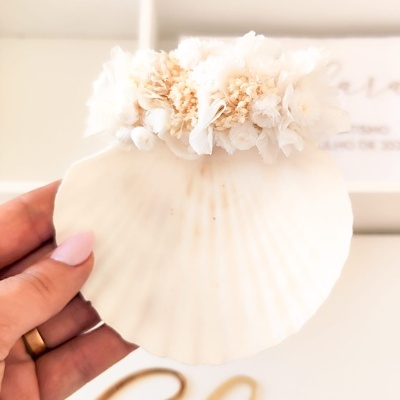 Concha branca decorativa com flores secas em tons creme, segurada por mão com unhas rosa e anel dourado