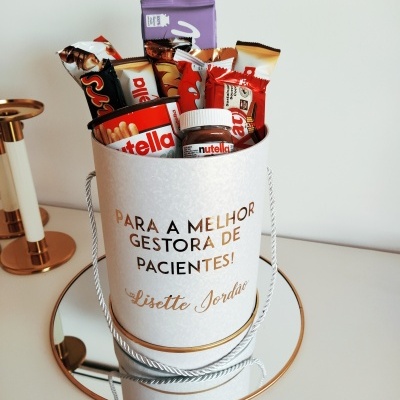 Saco branco com texto dourado e produtos Nutella e chocolates dentro, em cima de superfície branca com espelho redondo e castiçal ao lado.