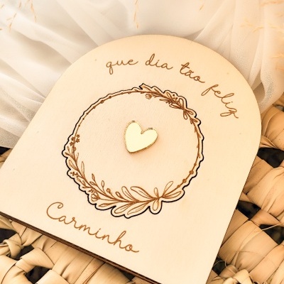 Placa decorativa de madeira com coração dourado e texto gravado, sobre fundo de tecido branco e cesta de palha