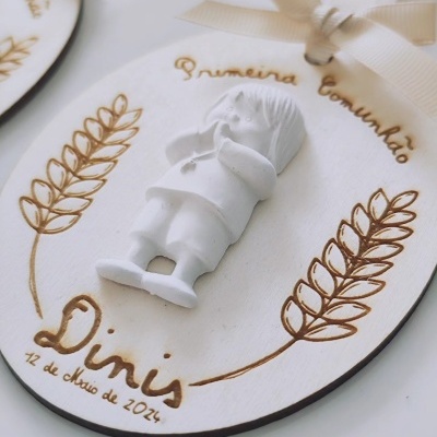 Plaqueta oval branca com figura de menino e texto dourado sobre primeira comunhão