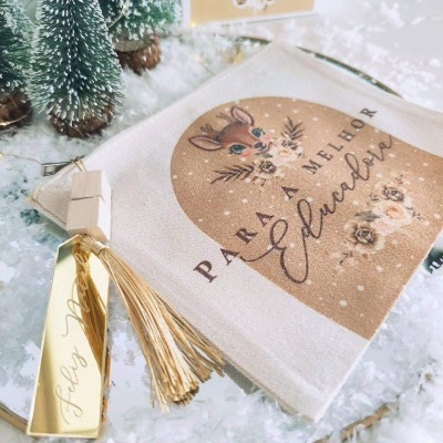 Saco tecido bege com estampa de veado e flores e texto para melhor educadora, etiqueta dourada félicitando Natal, mini árvores Natal e neve artificial