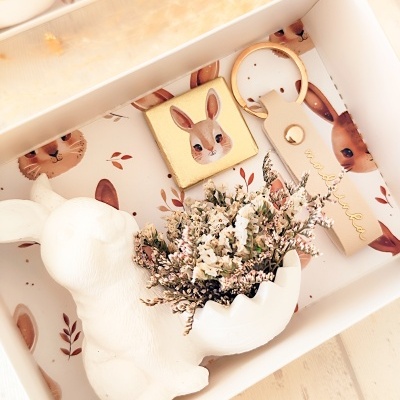 Conjunto com figura de coelho cerâmico, chaveiro de couro bege e cubo decorativo de coelho dentro de caixa branca com ilustrações.