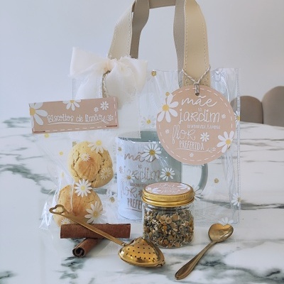Conjunto de chá com caneca azul, biscoitos, frasco com camomila, colher e infusor, em saco transparente com alças e laço branco