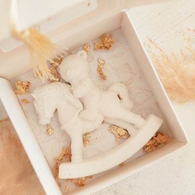 Escultura branca de ursinho num cavalo de balanço dentro de caixa branca com folha dourada.