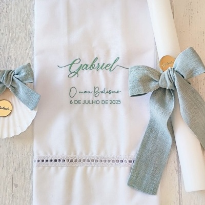 Toalha branca com bordado verde, rolo branco e concha decorados com laços verdes num fundo de madeira clara