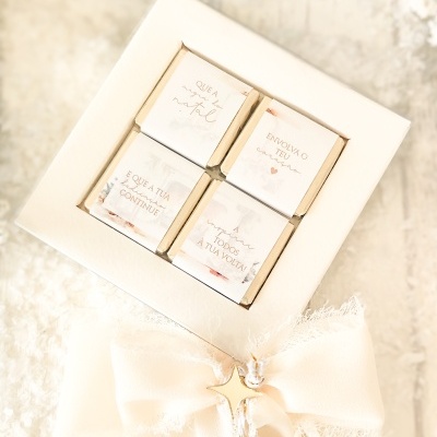 Caixa branca com quatro chocolates pequenos embrulhados em papel creme com frases em português e decorada com laço creme e estrela dourada.