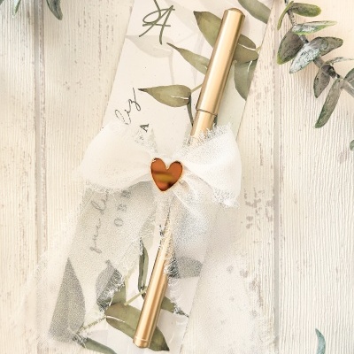 Caneta dourada com laço branco e cartão decorativo com folhas verdes