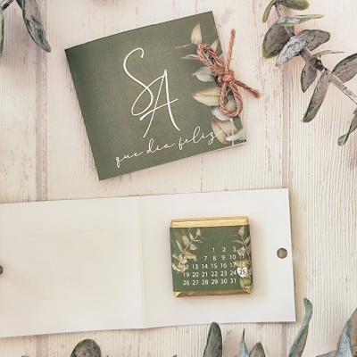 Calendário de chocolate com embalagem e cartão verde com texto, com ramos de folhas verdes ao redor numa superfície de madeira clara