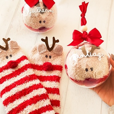 Meias peludas com padrão de riscas vermelhas e creme e decoração de rena dentro de bolinhas transparentes com nomes.