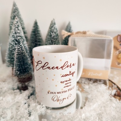 Caneca branca decorativa com texto em castanho, acompanhada de miniaturas de árvores de Natal e neve falsa