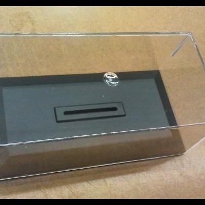 Caixa transparente retangular com base preta e tampa removível em superfície castanha.