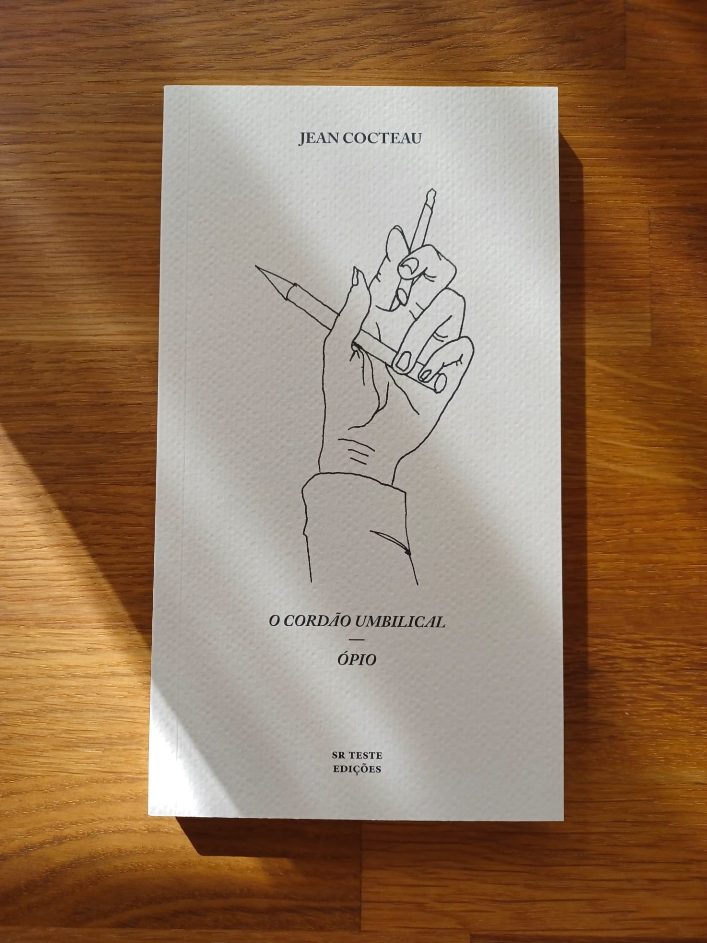 O Cordão Umbilical – Ópio, de Jean Cocteau Livro branco com desenho de mão segurando lápis e cigarro sobre mesa de madeira