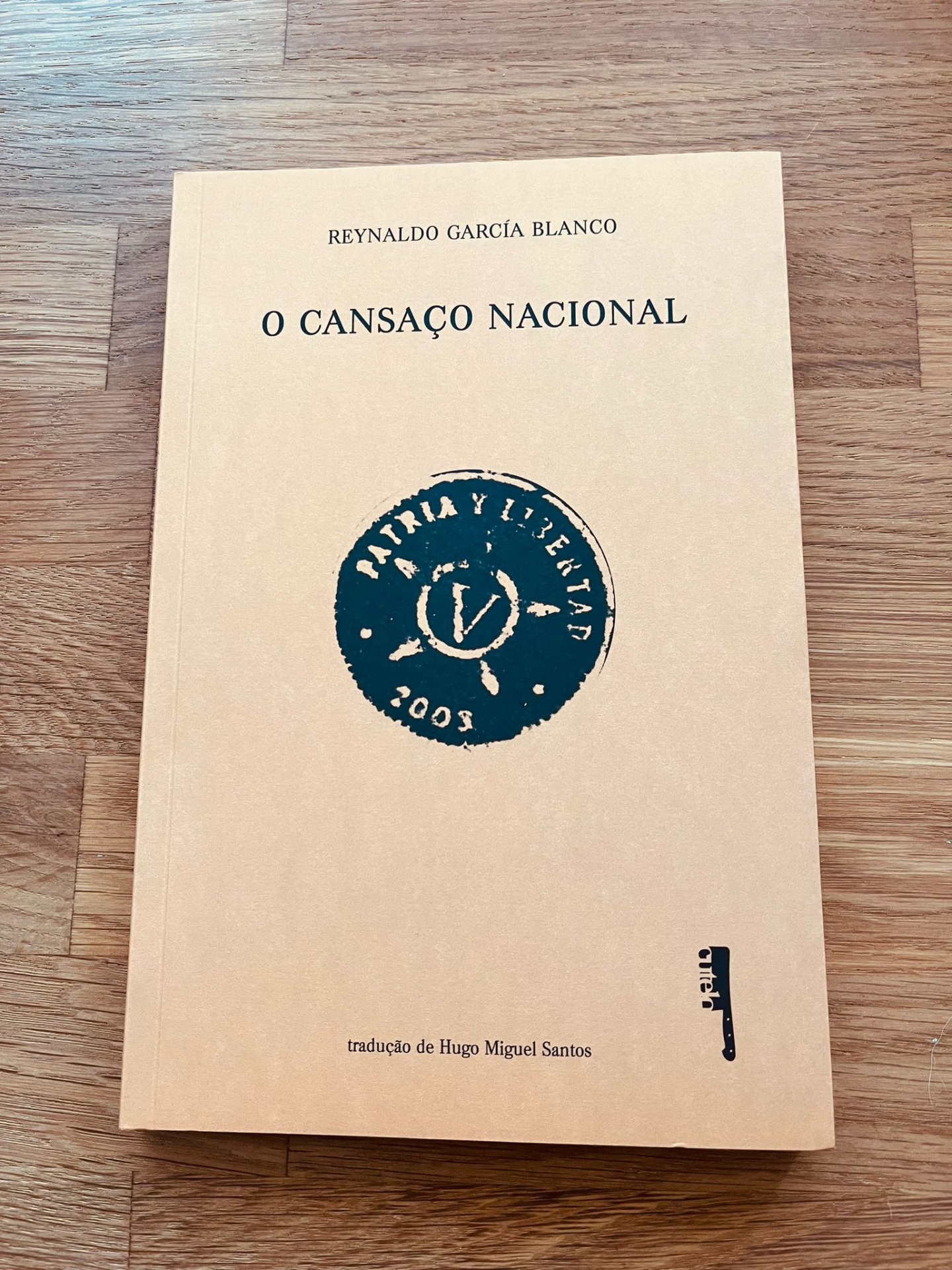 O Cansaço Nacional, de Reynaldo García Blanco Capa de livro amarelo claro com texto e selo azul escuro sobre mesa de madeira clara