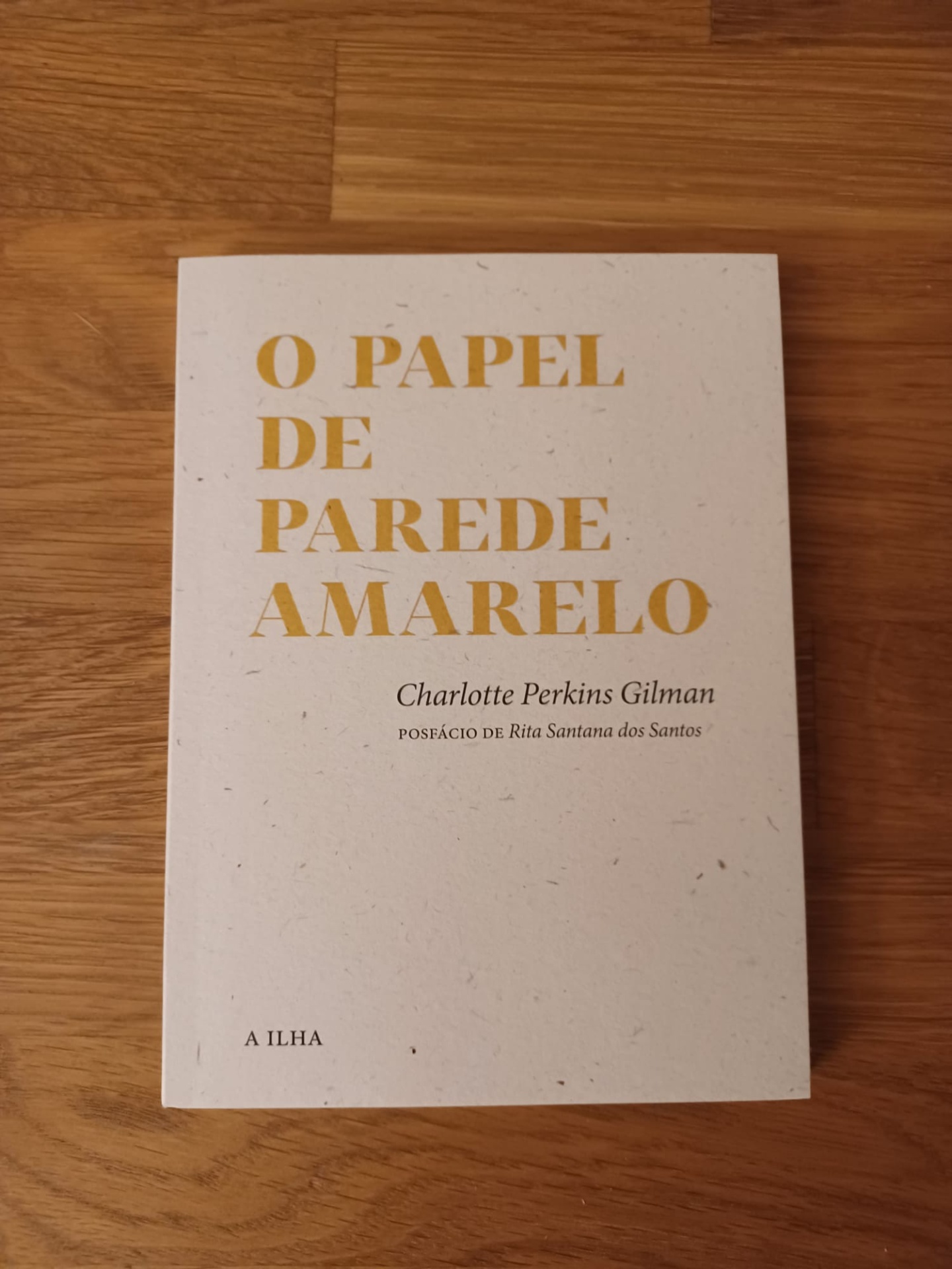 Papel de parede amarelo, de Charlotte Perkins Gilman Capa de livro branco com texto amarelo e preto sobre mesa de madeira
