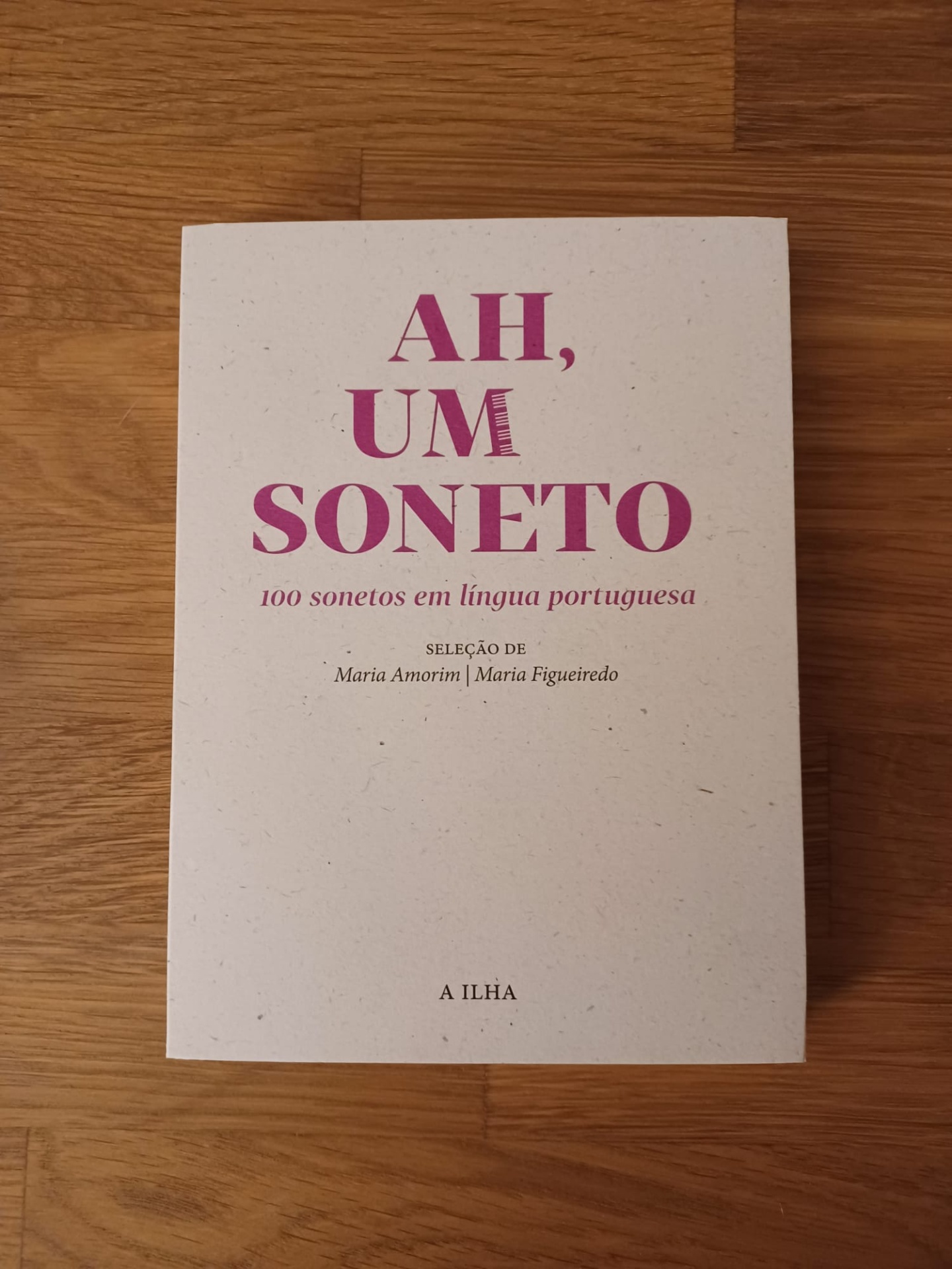Ah, um soneto - 100 sonetos em língua portuguesa Capa de livro branco com título e texto em rosa e preto sobre mesa de madeira clara