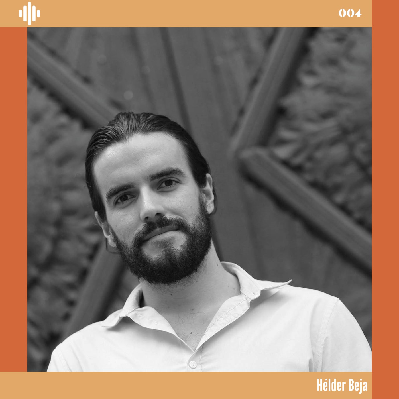 Homem com barba e cabelo escuros vestido com camisa branca em fundo de madeira e pedra