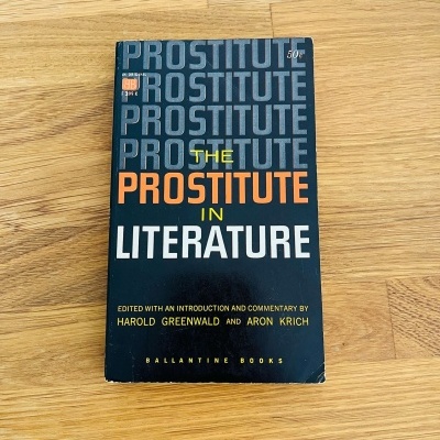 Livro 'The Prostitute in Literature' com capa preta e texto colorido sobre madeira clara
