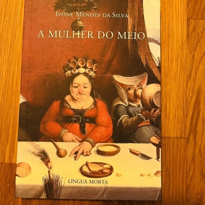 Capa do livro A Mulher do Meio com ilustração de duas figuras sentadas à mesa