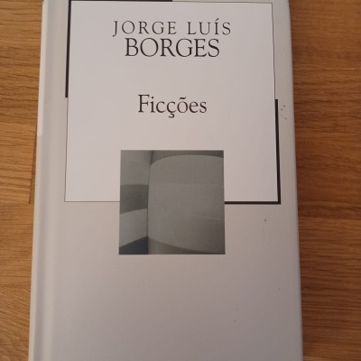 Livro Ficções de Jorge Luís Borges com capa branca e imagem abstrata cinza sobre mesa de madeira