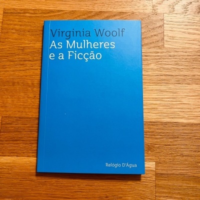 Capa azul do livro 'As Mulheres e a Ficção' de Virginia Woolf sobre madeira