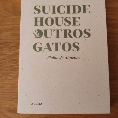 Capa de livro branco com texto verde 'SUICIDE HOUSE & OUTROS GATOS' e preto 'Fialho de Almeida', colocado numa mesa de madeira.