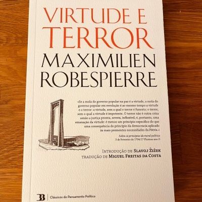 Capa de livro 'VIRTUDE E TERROR' com ilustração de guilhotina sobre mesa de madeira