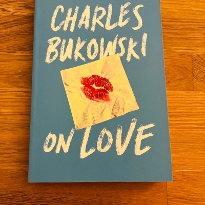 Livro 'CHARLES BUKOWSKI ON LOVE' com capa azul e imagem de beijo vermelho