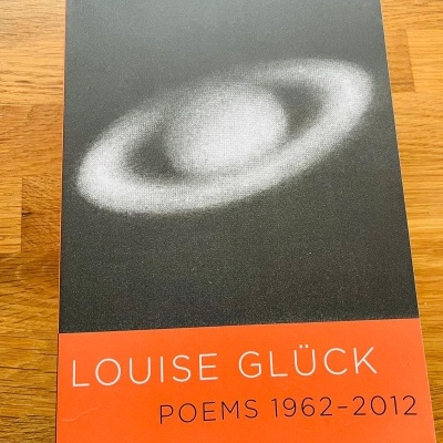 Livro com capa preta e vermelha com padrão branco em anel, texto LOUIS GLÜCK POEMS 1962-2012