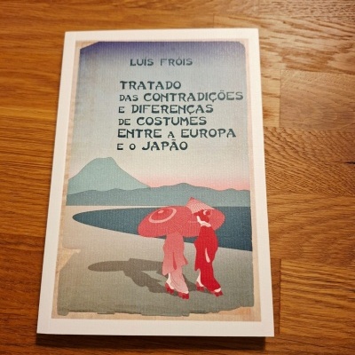 Capa de livro com ilustração de duas mulheres de quimono e sombrinhas vermelhas, título e autor em letra preta, sobre mesa de madeira.