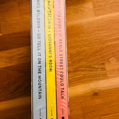 Três livros coloridos de James Baldwin alinhados verticalmente em chão de madeira