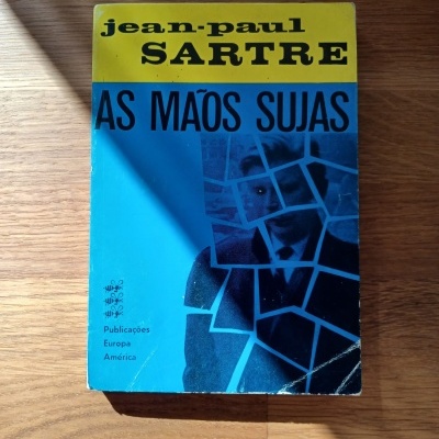 Capa do livro As Mãos Sujas de Jean-Paul Sartre com fundo azul e amarelo, sobre mesa de madeira.