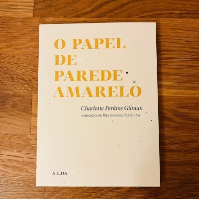 Livro 'O PAPEL DE PAREDE AMARELO' com capa branca e texto amarelo e preto sobre madeira