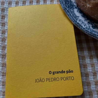 Caderno amarelo com texto 'O grande pão JOÃO PEDRO PORTO' ao lado de prato com pão sobre toalha quadriculada