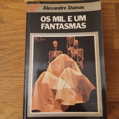Livro 'OS MIL E UM FANTASMAS' de Alexandre Dumas com capa que mostra esqueletos e tecido sobre fundo preto.