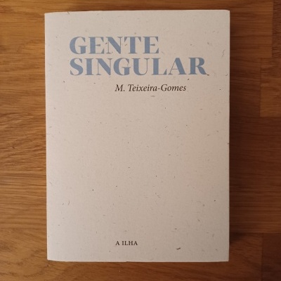 Livro GENTE SINGULAR de M. Teixeira-Gomes com capa branca e texto azul sobre mesa de madeira