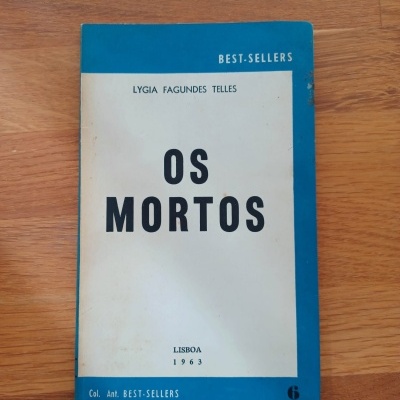Livro 'Os Mortos' de Lygia Fagundes Telles com capa branca e azul sobre superfície de madeira clara
