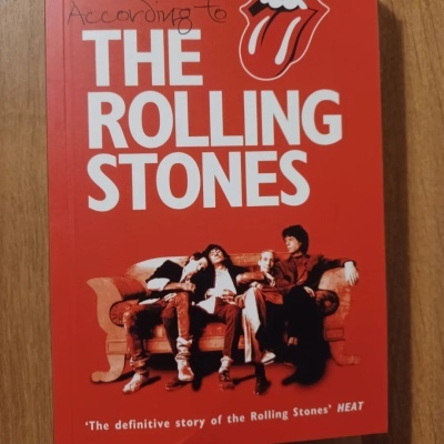 Capa de livro vermelha com texto branco THE ROLLING STONES e imagem da banda sentada em sofá