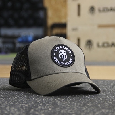 Boné verde-oliva com emblema redondo branco e preto, sobre tapete de ginásio com caixas ao fundo