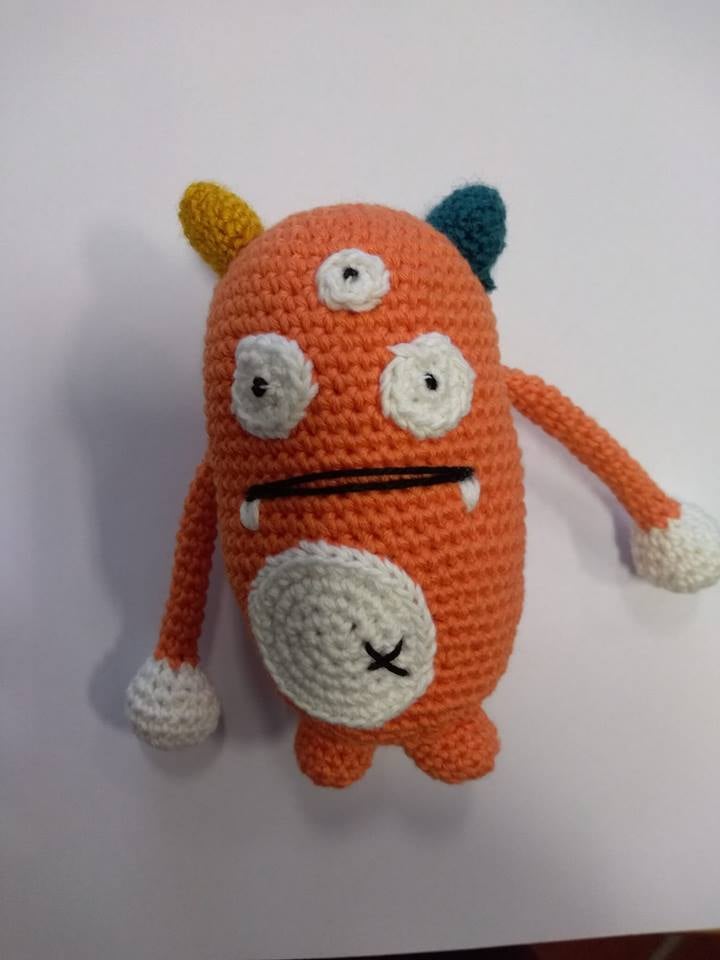 Boneco de tricot laranja com olhos e detalhes brancos, e chumaços coloridos no topo
