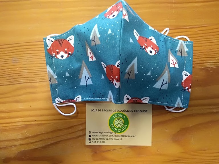 Máscara de tecido azul com estampa de raposas e pinheiros sobre mesa de madeira, com cartão de loja ecológica.