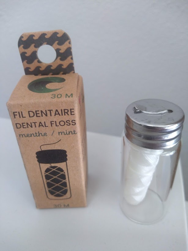 Embalagem castanha clara de fio dentário com texto FIL DENTAIRE DENTAL FLOSS e recipiente transparente com tampa prateada contendo fio dentário