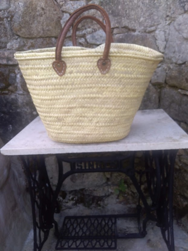 Sacola de palha com pegas de couro sobre mesa de ferro com tampo branco frente a parede de pedra