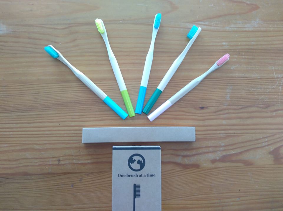 Cinco escovas de dentes com cabos brancos e cores diversas, caixa de papelão com frase 'One brush at a time', sobre superfície de madeira.