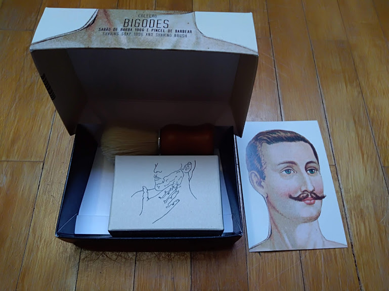 Caixa aberta com pincel, bloco de papel e cartão com ilustração de homem com bigode sobre superfície de madeira.